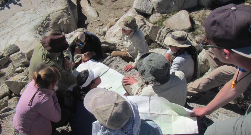 A group of people sit on a rocky surface while examining maps